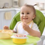 Cute little baby eating food in high chair at kitchen